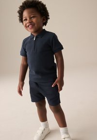 Polo bleu marine à manches courtes avec une fermeture éclair, assorti à un short de la même couleur. La tenue est complétée par des baskets blanches et des chaussettes à motifs.