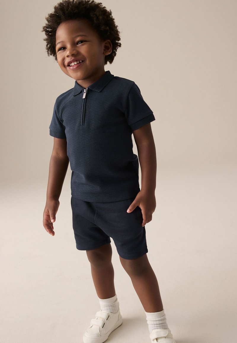 Polo bleu marine à manches courtes avec une fermeture éclair, assorti à un short de la même couleur. La tenue est complétée par des baskets blanches et des chaussettes à motifs.