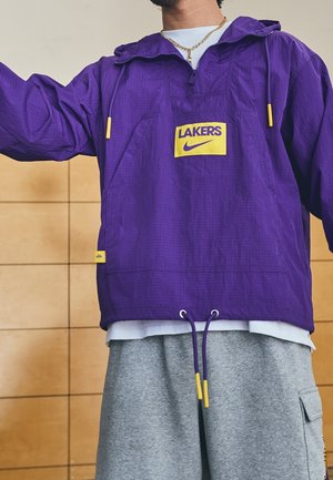 Persoon die een paarse Lakers windjack met gele accenten en grijze joggingbroek draagt, staat tegen een beige betegelde muur.