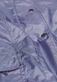 Leichte lila Jacke mit einer glänzenden, strukturierten Oberfläche, ausgestattet mit schwarzen Druckknöpfen und sichtbaren Nähdetails auf dem Stoff.