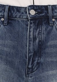 Tmavomodré džínsy z denimu s hladkou textúrou, s kovovou prieraznou väzbou a pútkami na opasok. Obsahujú jemné vyblednutie a kontrastné stehy.