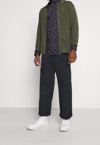 Man i vita sneakers, mörka byxor med uppvikt nederkant, blommönstrad marinblå skjorta och olivgrön zip-up jacka mot en enfärgad bakgrund.