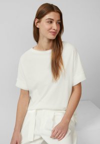 Biała koszulka z krótkim rękawem z miękkiego materiału, z okrągłym dekoltem i podwiniętymi mankietami, w zestawieniu z jasnymi, dopasowanymi spodniami.