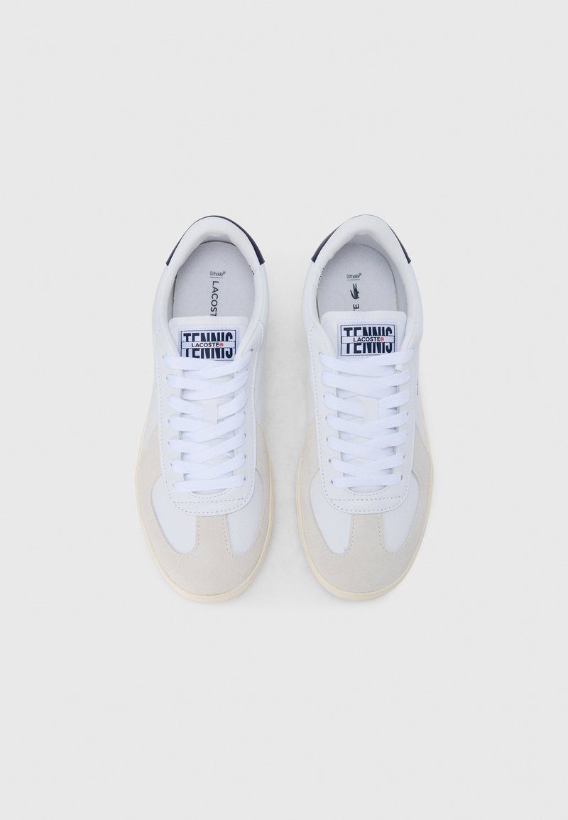 Par hvide og beige Lacoste tennissko med hvide snørebånd og mærkning på tungen, set oppefra på en ensfarvet baggrund.