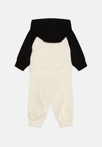 Ensemble de sweat à capuche en noir et crème. Doté de manches longues, de poignets élastiques et d’un pantalon crème assorti avec une taille élastique. Tissu doux.