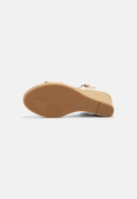 Chaussure à talon compensé couleur beige avec semelle en caoutchouc texturée et boucle à la cheville, vue par le dessous sur fond blanc.