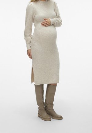 Femme enceinte portant une robe en maille beige à manches longues avec une fente latérale et des bottes taupe montant jusqu'aux genoux, main reposant sur le ventre.