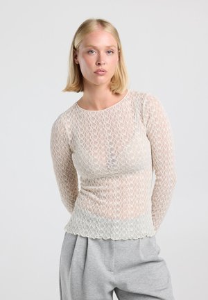 Vrouw met steil blond haar draagt een doorschijnende, lange mouwen kanten top en lichtgrijze broek, staand tegen een effen achtergrond.