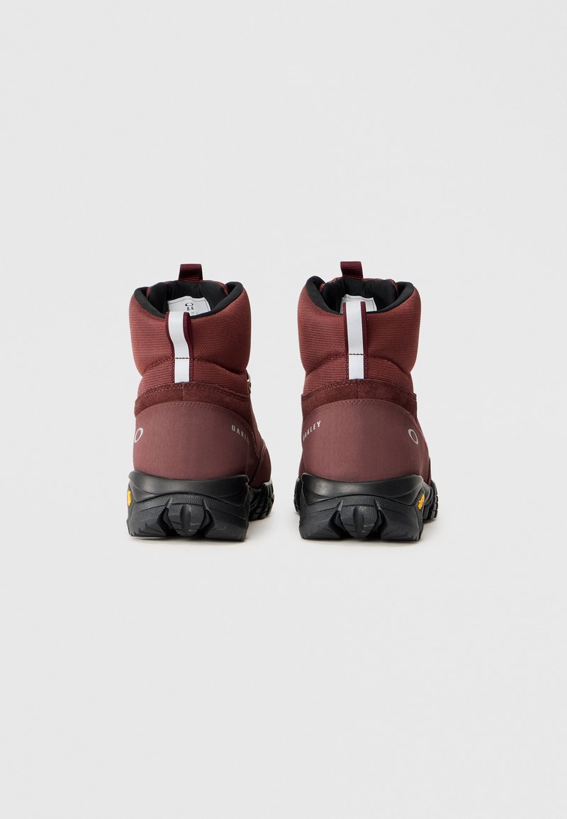 Botas de senderismo color burdeos con un collar acolchado, suela negra y tacos prominentes. Incluye una pestaña blanca para tirar y detalles de marca en los lados.