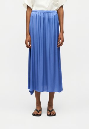 Osoba ubrana w luźną niebieską spódnicę sięgającą kostek, biały top i czarne sandały, stojąca na tle jednolitego jasnego tła.