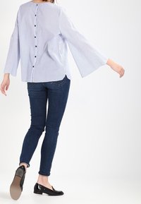 Camicia a righe blu chiaro e bianca con scollatura rotonda, maniche a campana e bottoni neri sul retro; abbinata a jeans di denim blu scuro e scarpe nere.