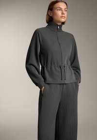 Hanorac gri cu fermoar și guler înalt, cu talie elastică și șnur reglabil, având o textură cu nervuri, asortat cu pantaloni largi de aceeași culoare.