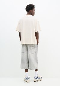 Beige pamut póló rövid ujjakkal, szürke denim culotte -kal párosítva, ami hátul logóval ellátott zsebet tartalmaz. Fehér és kék sportcipővel viselve.