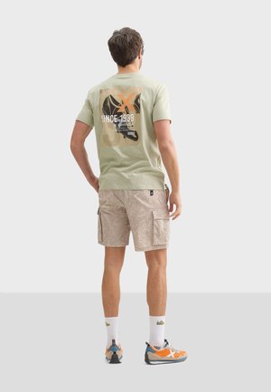 Camiseta de algodón verde claro con estampado gráfico en la espalda, combinada con pantalones cortos cargo beiges con estampado, calcetines blancos y zapatillas de colores.