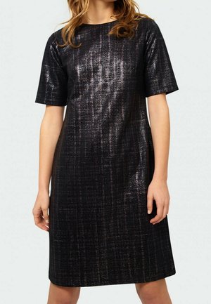 Frau trägt ein knielanges, kurzärmliges schwarzes Kleid mit einem dezenten glänzenden, strukturierten Muster vor hellem Hintergrund.