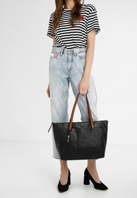 Sac cabas en cuir noir avec poignées en cuir marron, de forme rectangulaire, fermeture zippée et porte-clés; assorti avec une chemise rayée et un jean clair.