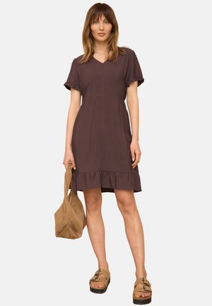 Femme portant une robe marron à manches courtes, tenant un sac à main beige, debout en sandales compensées beige sur un fond clair uni.