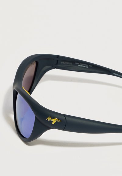 Ματ μαύρα γυαλιά ηλίου Maui Jim με φακούς μπλε απόχρωσης και χρυσό λογότυπο στο πλάι, εμφανίζονται σε ελαφρώς κεκλιμένη γωνία σε λευκό φόντο.