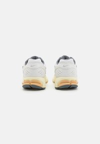 Nike sportcipő fehér hálós felsőrésszel, szürke részletekkel, narancssárga és bézs párnázott talppal, valamint geometrikus mintával a sarokon.