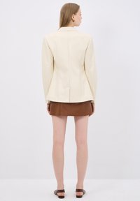 Blazer crema su misura con spalle strutturate e vita aderente, indossato sopra una minigonna in pelle marrone. La vista posteriore mette in risalto linee pulite.