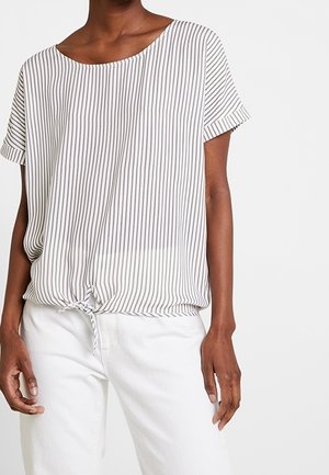 Frau trägt eine kurzärmelige weiße Bluse mit dünnen vertikalen schwarzen Streifen und einer Schleife am Saum, kombiniert mit weißen Hosen.