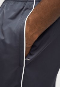 Σκούρο δέρμα χεριού τοποθετημένο μέσα στην πλαϊνή τσέπη σκούρων γκρι σορτς με λευκό περίγραμμα κατά μήκος της άκρης της τσέπης.