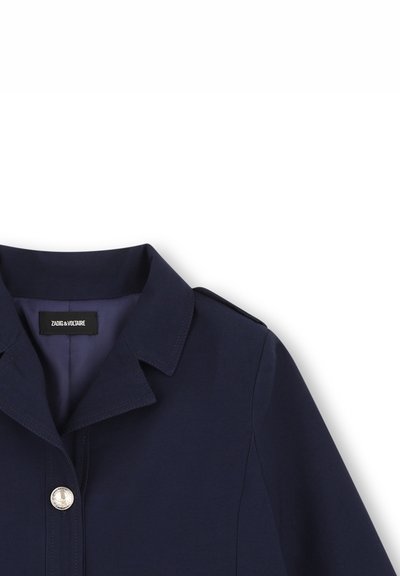 Veste bleu marine à col avec épaulette aux épaules et bouton argenté, partiellement visible sur un fond blanc.