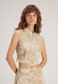 Top corto floral en beige claro con patrones florales en blanco y oro. Cuenta con un cuello y cierre de botones en el frente. Textura de lino suave.