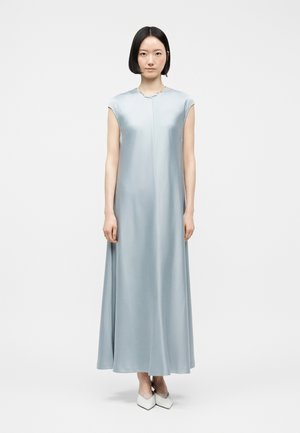 Donna in piedi, frontale, che indossa un abito azzurro chiaro in raso senza maniche, lungo fino al pavimento, e scarpe bianche con punta appuntita su uno sfondo bianco uniforme.