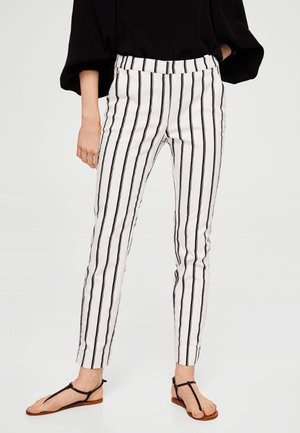 Kvinde iført hvide tætsiddende bukser med lodrette sorte striber, sort top med 3/4 ærmer og sorte flade sandaler.