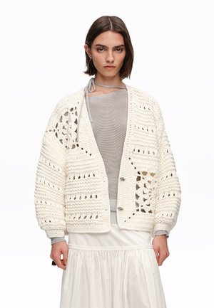 Mujer con un cárdigan blanco de ganchillo grueso sobre una camiseta gris clara y una falda blanca fluida, de pie frente a un fondo blanco liso.