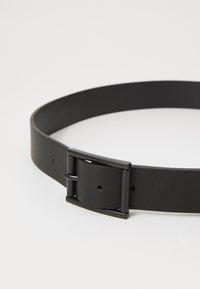 Černý kožený pásek s matným povrchem, obdélníkovou kovovou sponou a několika otvory pro nastavení velikosti. Jednoduchý a funkční design.