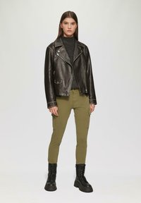 Zwarte leren bikerjack met zilverkleurige hardware, olijfgroene cargo broek en zwarte stevige combatboots. Model staat tegen een lichte achtergrond.