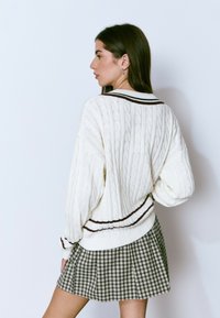 Joven mujer con un suéter blanco de punto trenzado con rayas oscuras y una falda plisada a cuadros verdes, de pie contra un fondo blanco.