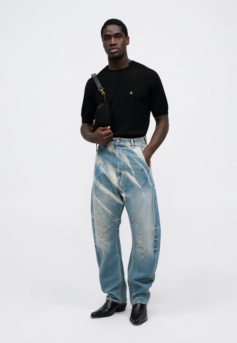 Maglietta nera a maniche corte in maglia, jeans baggy azzurri chiari con dettagli di lavaggio, e stivali neri a punta. Il modello posa con una borsa a tracolla nera.