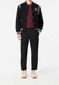 Svart ulljacka med crèmefärgade ränder och Lacoste-logotyp, bordeauxfärgad t-shirt, svarta elastiska byxor och ljusgrå sneakers med orange accenter.