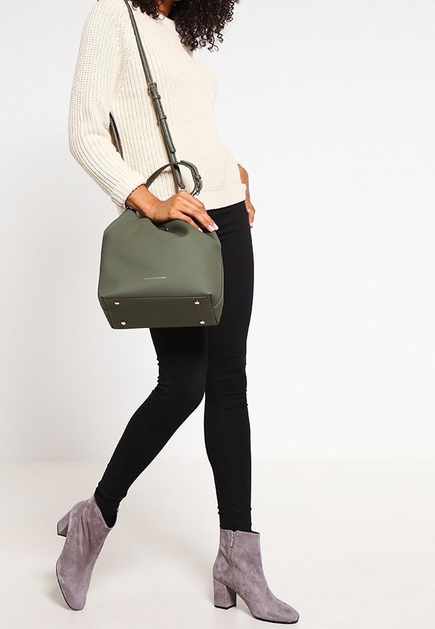 Femme portant un pull crème, des leggings noirs, des bottines en daim gris et portant un sac à main vert olive avec une bandoulière.