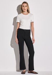 Pantaloni neri a zampa d'elefante con una texture liscia e due bottoni frontali, abbinati a una t-shirt bianca aderente a maniche corte.