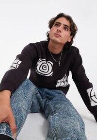 Suéter negro con espiral blanca y diseños abstractos, combinado con jeans de denim azul desgastado. Tela natural, corte relajado, detalles texturizados.