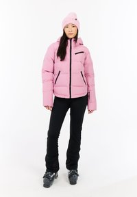 Ružová pufová bunda so zipsom vpredu, čiernymi akcentmi a kapucňou. Čierne priliehavé nohavice a lyžiarske topánky dotvárajú zimný outfit.