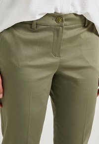 Pantalons ajustés vert olive en tissu lisse, avec une fermeture éclair, un bouton, des poches avant et des plis subtils le long des jambes.