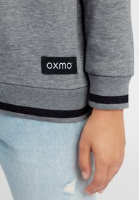 Persona que lleva jeans rasgados de color azul claro y un sudadera gris con rayas negras y una etiqueta "oxmo" en el dobladillo.