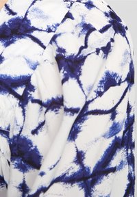 Tissu blanc avec un motif abstrait bleu, comprenant des éclaboussures et des traits. La texture semble lisse avec des détails subtils de plissement.