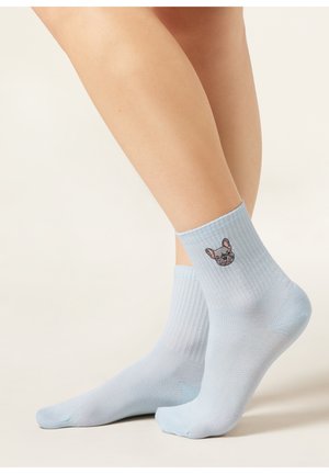 Jambes portant des chaussettes bleu clair côtelées avec un visage de bouledogue français brodé sur le côté extérieur de la chaussette droite.