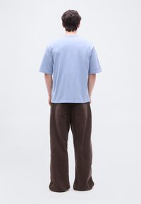 Άτομο που στέκεται με την πλάτη στην κάμερα, φορώντας ανοιχτό μπλε κοντομάνικο πουκάμισο και χαλαρό καφέ παντελόνι, μπροστά σε απλό λευκό φόντο.