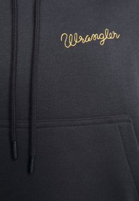 Juodas megztinis su švelnia medžiaga, su kengūros kišenių, užtrauktuku reguliuojamu kapuce ir aukso siuvinėtu "Wrangler" logotipu ant krūtinės.