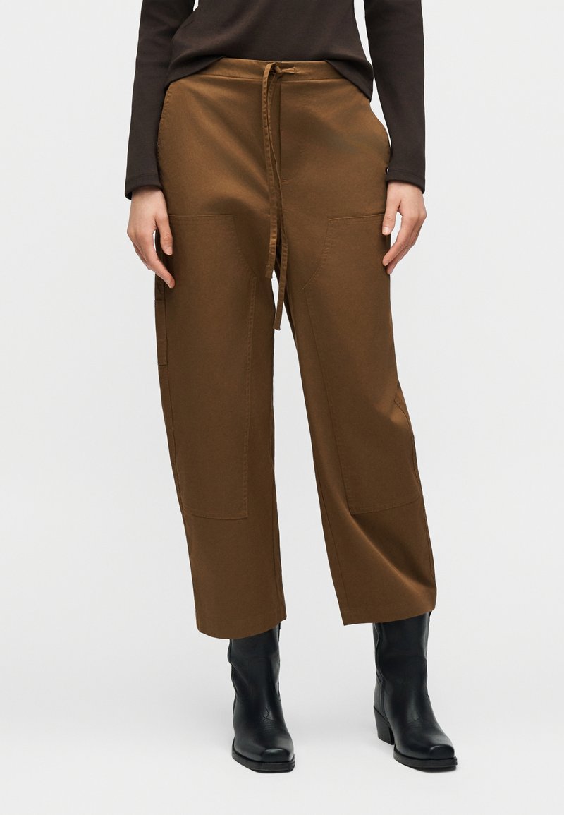 Καφέ παντελόνι φαρδιάς γραμμής από βαμβακερό μείγμα με κορδόνι στη μέση, διαθέτει δύο μεγάλες μπροστινές τσέπες και λείο ύφασμα. Ορατές μαύρες μπότες αστραγάλου.