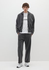 Grauer Fleece-Zip-Hoodie mit Fronttaschen, kombiniert mit passenden grauen Jogginghosen. Ein weißes T-Shirt darunter hat den Text "NEW YORK". Weiße Sneakers.