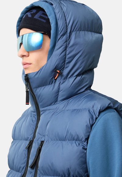 Blauer Steppwesten mit hohem Kragen und Kapuze, ausgestattet mit einem schwarzen Reißverschluss und orangefarbener Zuglasche. Tragen mit einer schwarzen Mütze und reflektierenden Sonnenbrillen.