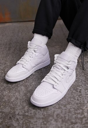 Baskets montantes blanches à lacets portées avec des chaussettes blanches et un pantalon noir sur un sol gris texturé.
